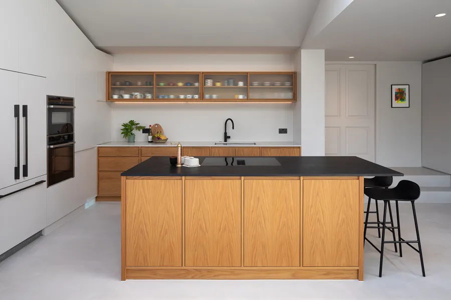 Oak veneered kitchen island with dark worktop and hob, sink run and glazed wall units behind.