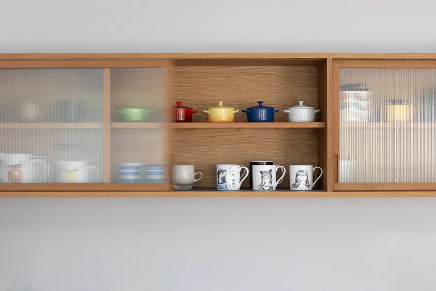 Oak wall unit with a reeded glass door slid part open to reveal coloured pots and mugs.