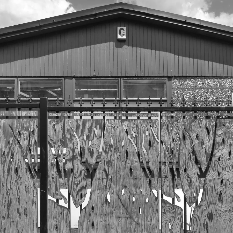 Black and white photo of the workshop exterior behind a fence of cut plywood panels.